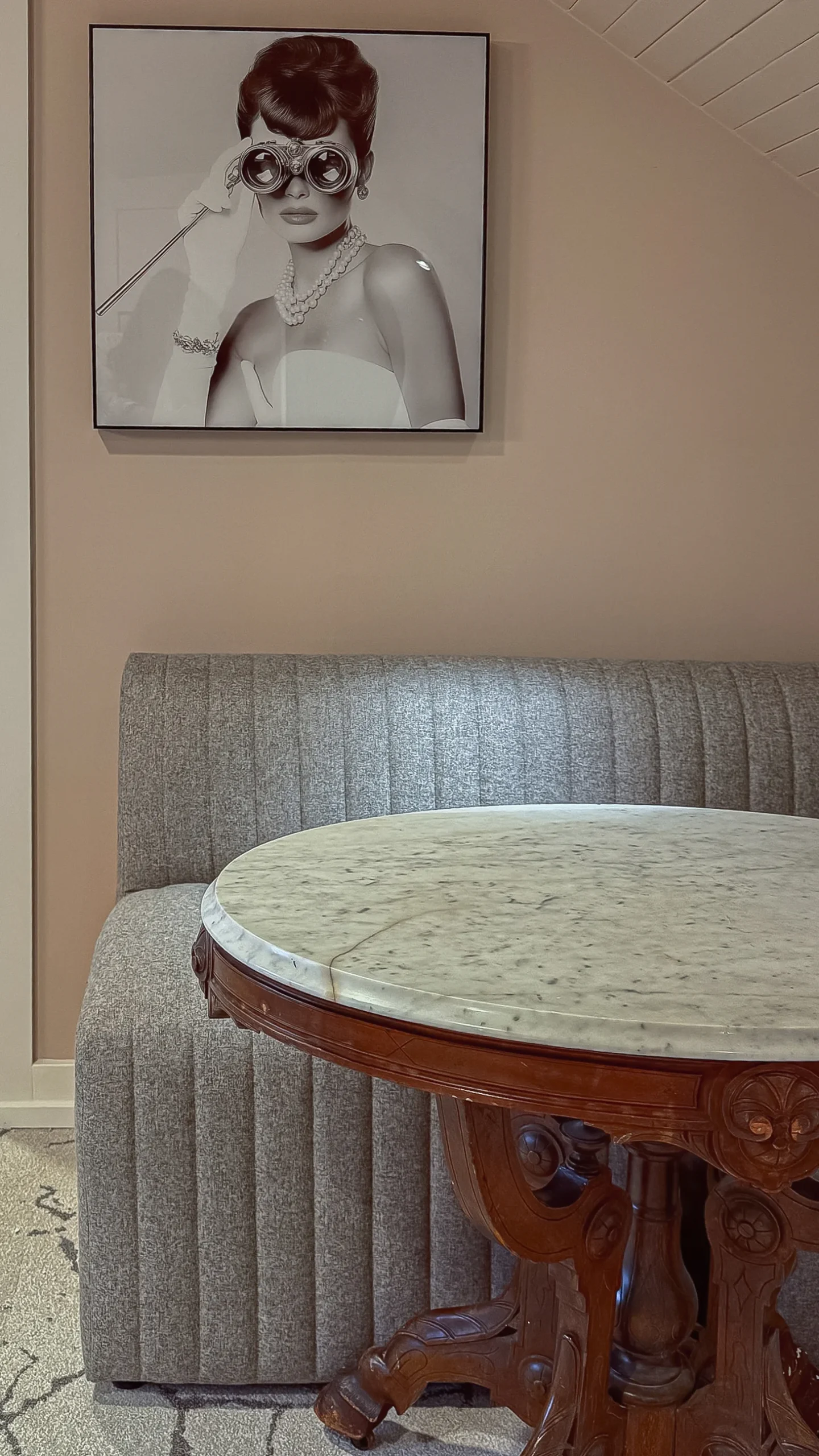 Close-up vertical shot of a seating area featuring a small round table with a marble top and a carved dark wood base, next to a fluted gray fabric bench. A black and white poster of a woman in binoculars is on the wall.