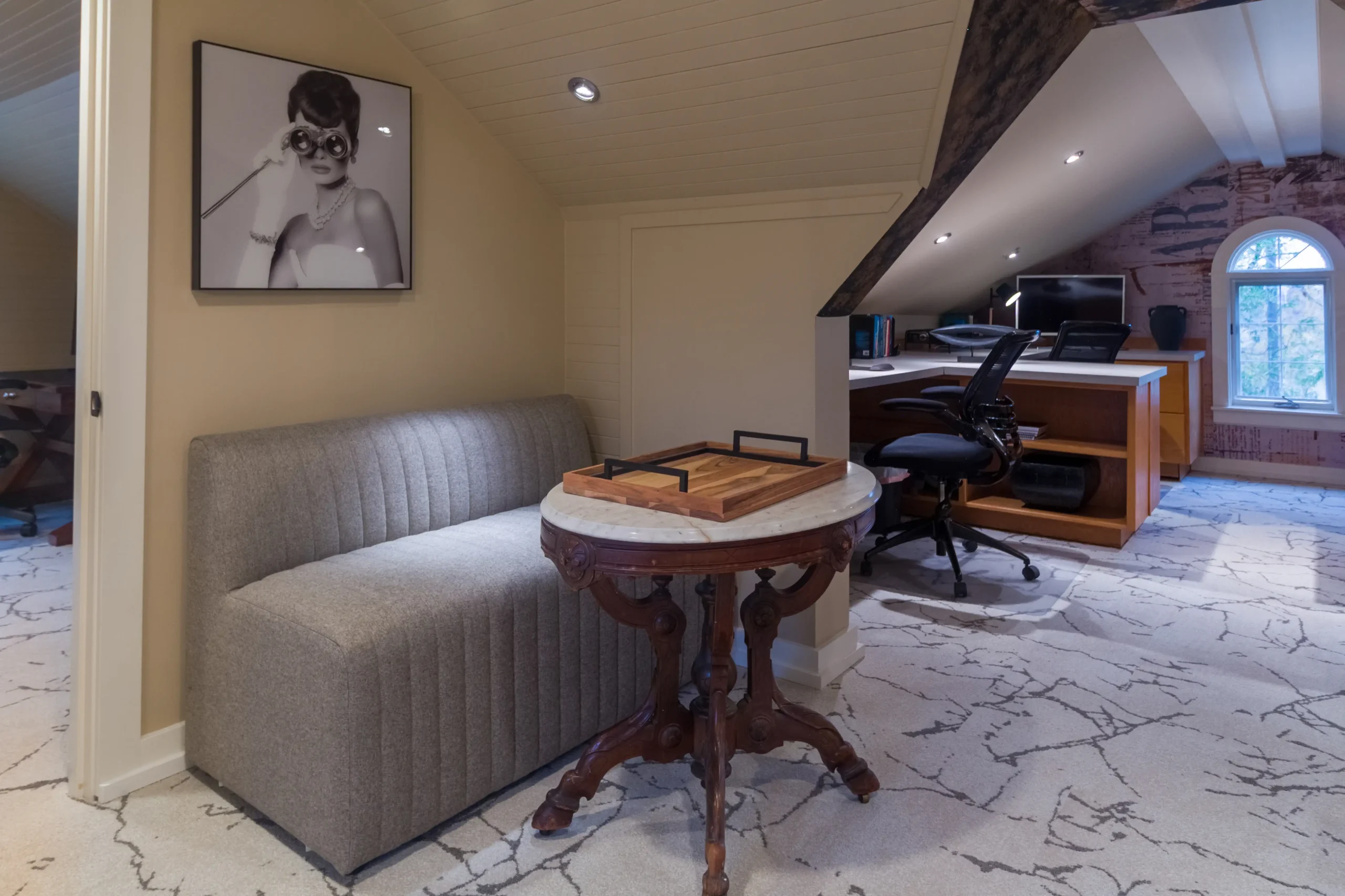Office lounge area featuring a gray fluted bench, marble-topped antique-style table, and a black and white framed print of a woman. A coworking office workspace is visible behind the seating area.