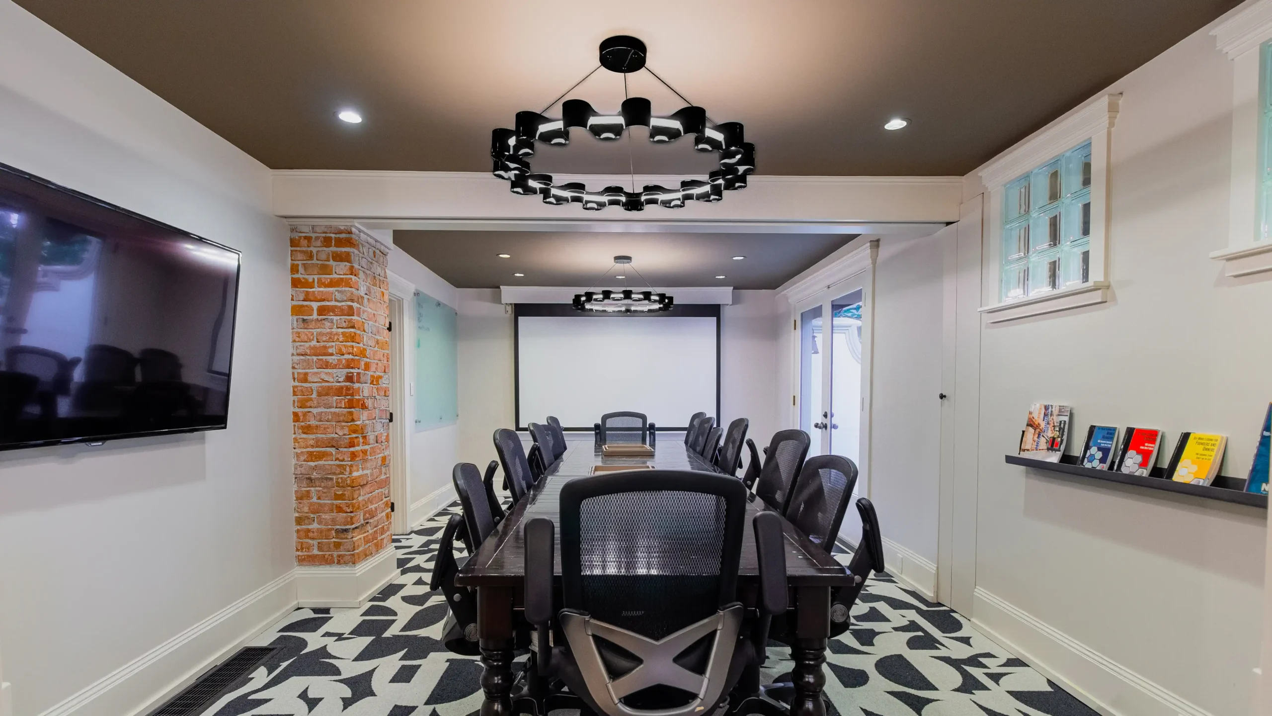 Wide, modern conference room with a large dark table surrounded by mesh-backed chairs, an exposed brick column, and a stylish black ring chandelier. The room features a screen projector and geometric carpet.