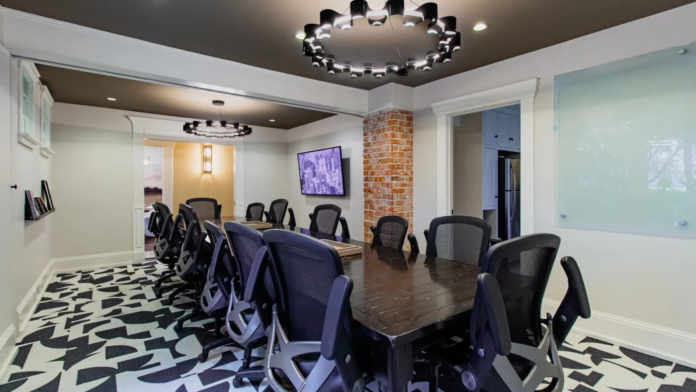 Wide view of a modern conference room setup with a long dark table, black mesh chairs, and a distinct black and white geometric carpet. A TV screen, brick column, and glass whiteboard are visible.