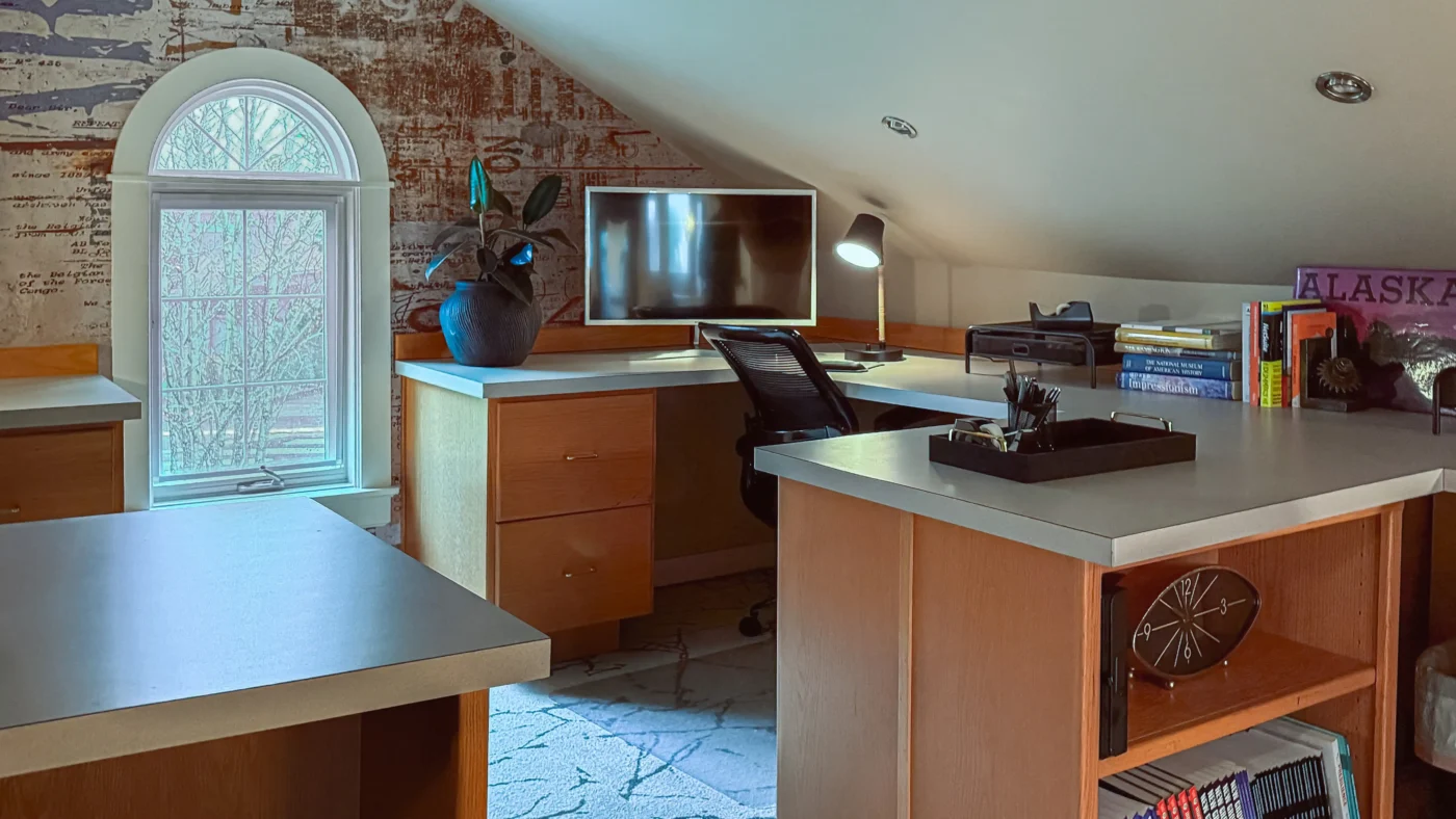 Coworking office space in an attic-style room with sloped ceilings, featuring a large desk area with light wood cabinets, computer monitors, and an arched window on a wall with expressive wallpaper.