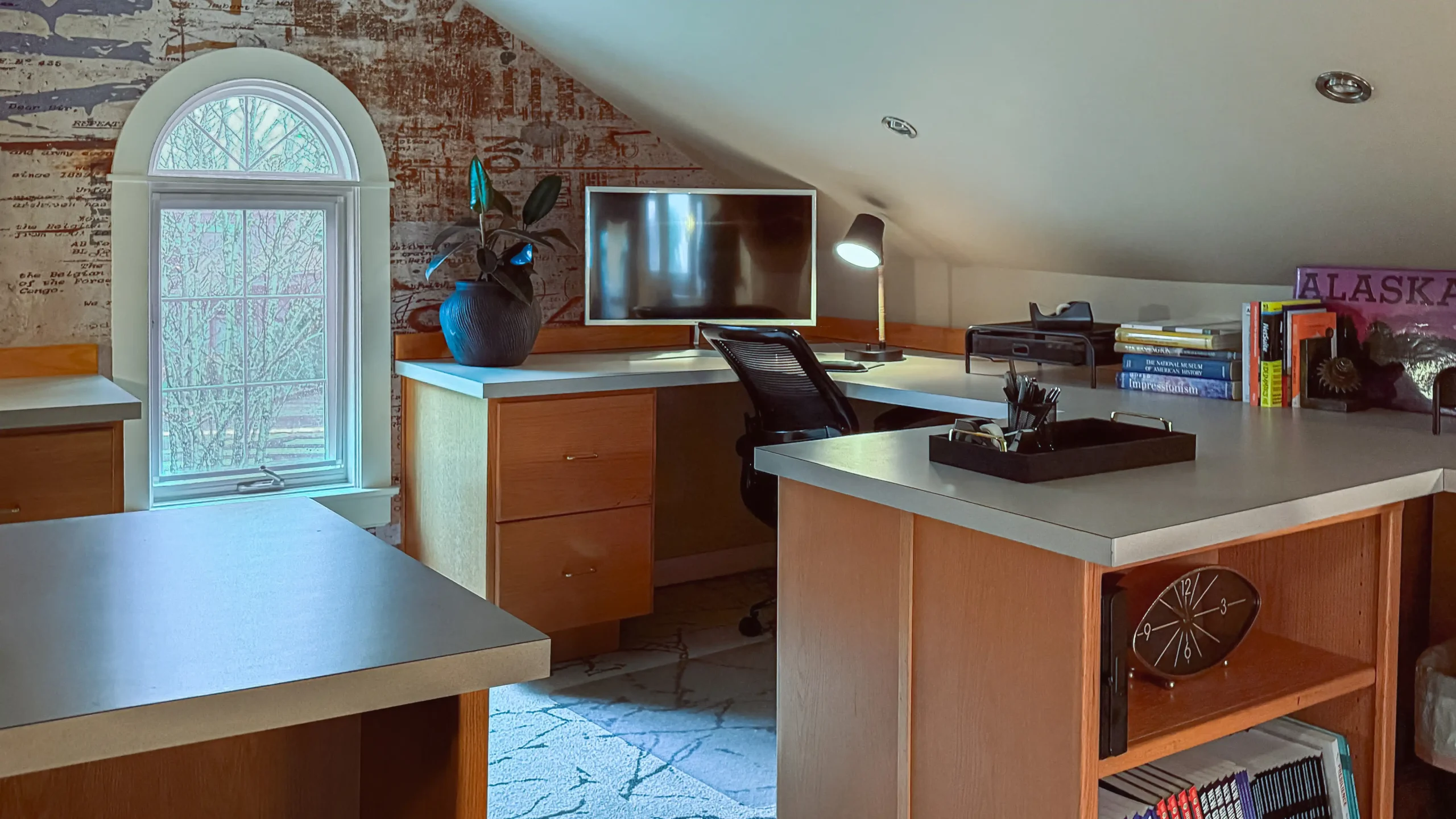 Coworking office space in an attic-style room with sloped ceilings, featuring a large desk area with light wood cabinets, computer monitors, and an arched window on a wall with expressive wallpaper.