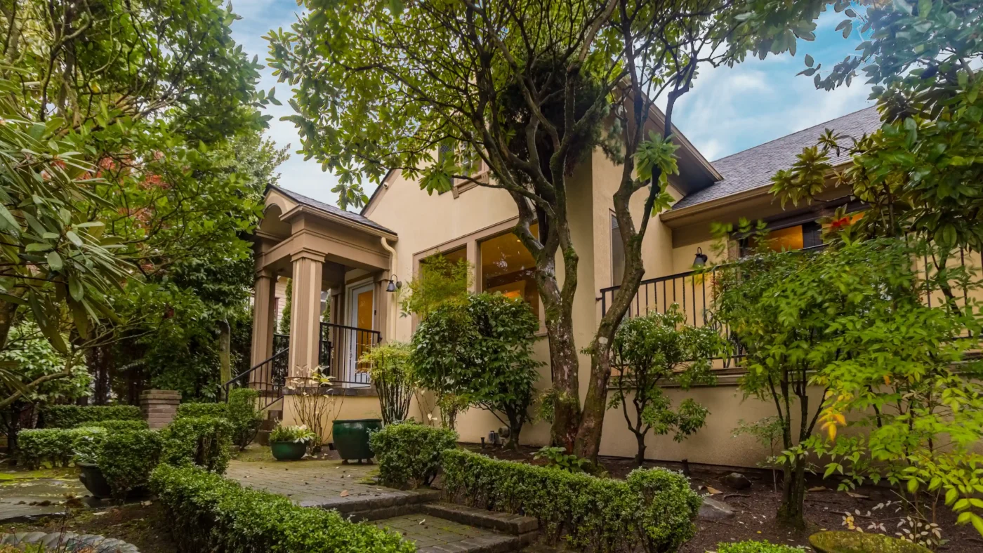 Exterior view of a light-colored stucco building with a pillared entrance, surrounded by lush green trees, manicured hedges, and a tiered stone pathway.