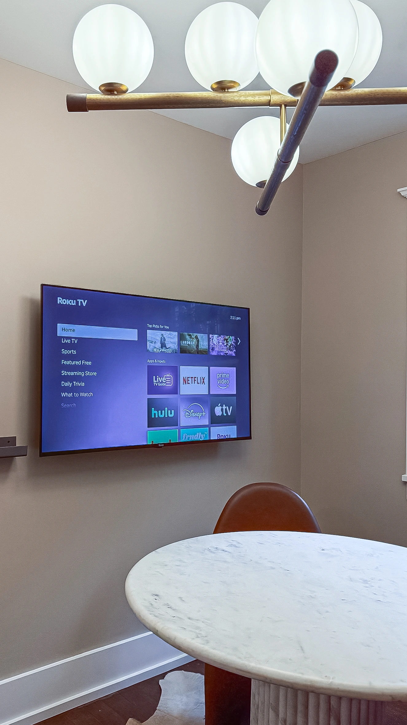 Vertical shot of a small meeting room showing a wall-mounted TV displaying the Roku TV home screen above a round marble table and a brown leather chair.