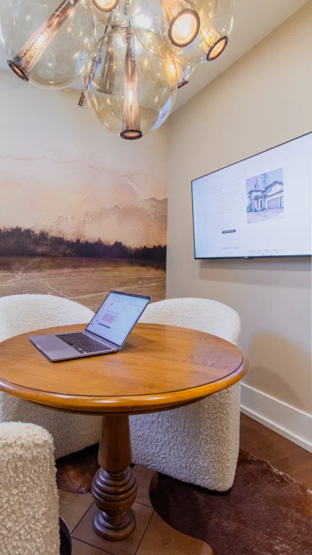 Vertical close-up of a laptop open on a round wood meeting table, with a wall-mounted TV on the right and a globular chandelier above.