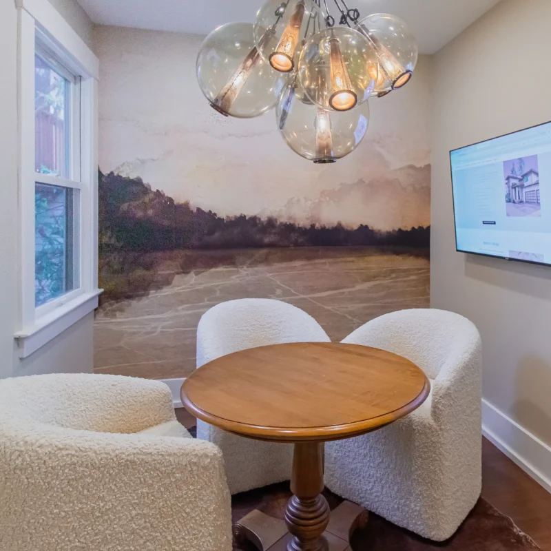Wide view of a small meeting room with a round wood table, three white sherpa-style chairs, a glass chandelier, and an abstract landscape wallpaper.