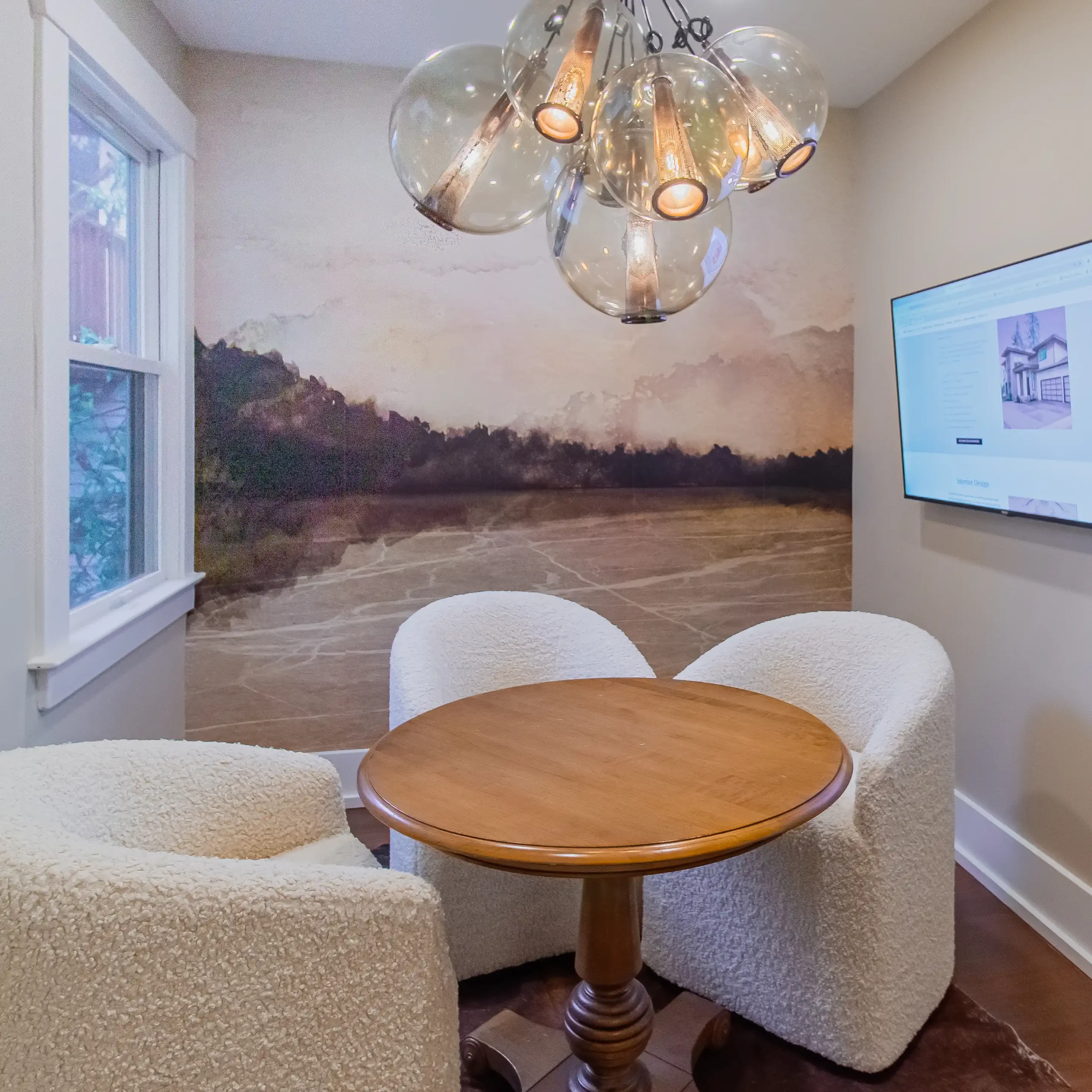 Wide view of a small meeting room with a round wood table, three white sherpa-style chairs, a glass chandelier, and an abstract landscape wallpaper.