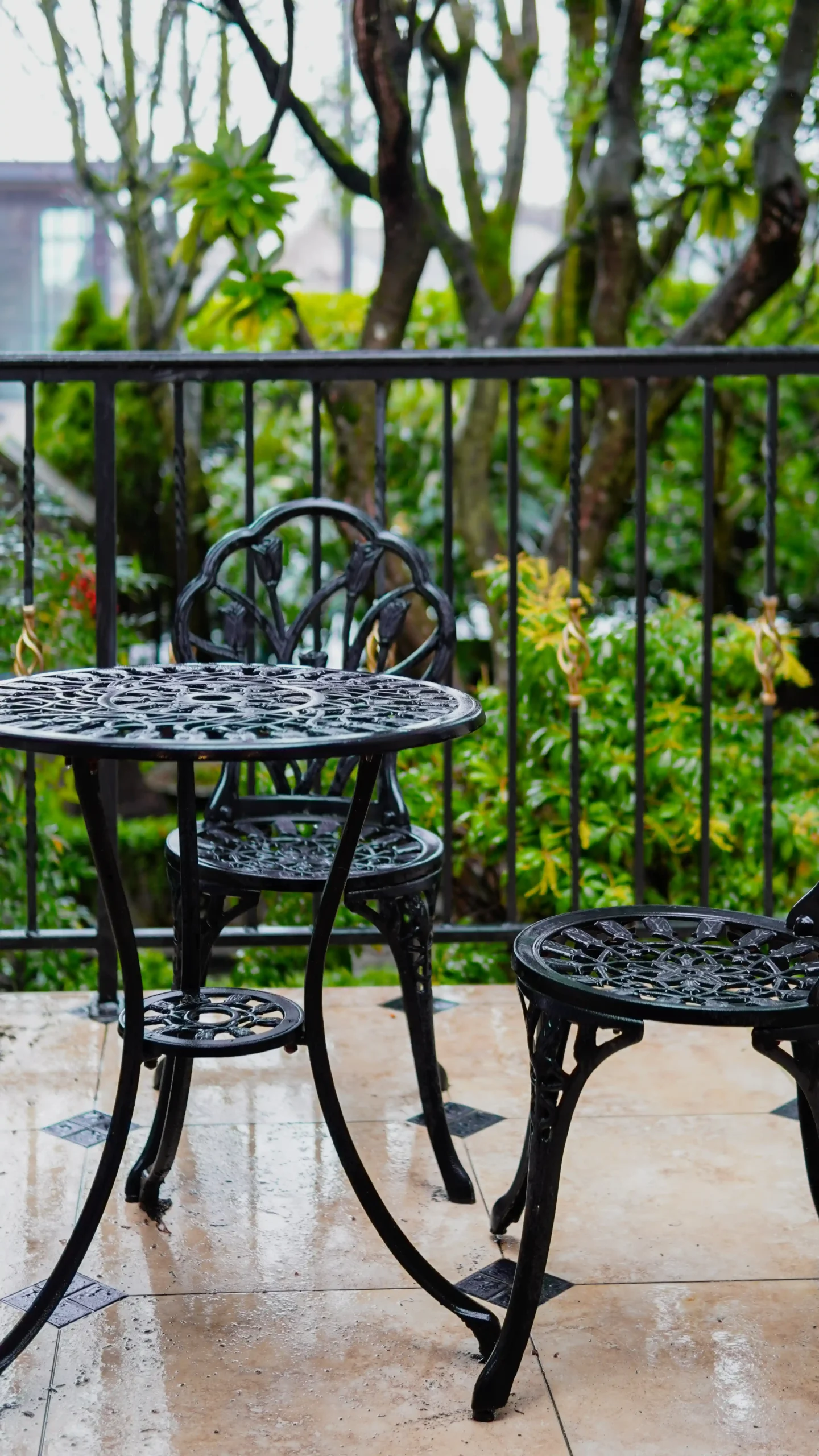 Close-up of a black, ornate cast iron outdoor bistro table and chairs on a patio with decorative tile and a black metal railing overlooking lush green trees.