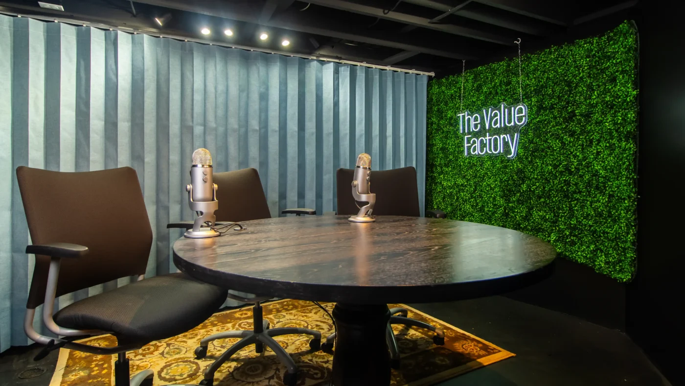 Podcast recording studio with a round black table, three office chairs, and two microphones on arms. Features a green hedge wall with a neon sign reading 'The Value Factory' and a blue backdrop curtain.
