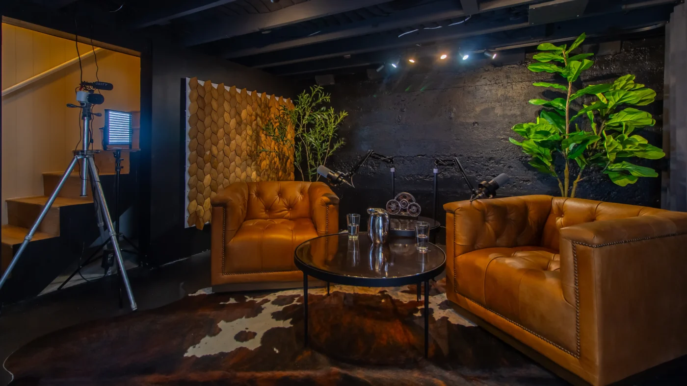 Podcast recording studio lounge area featuring two large, tufted brown leather armchairs, a round glass table, and two microphones on stands. The backdrop is a dark, textured wall with a fiddle leaf fig plant and a wooden scale pattern.