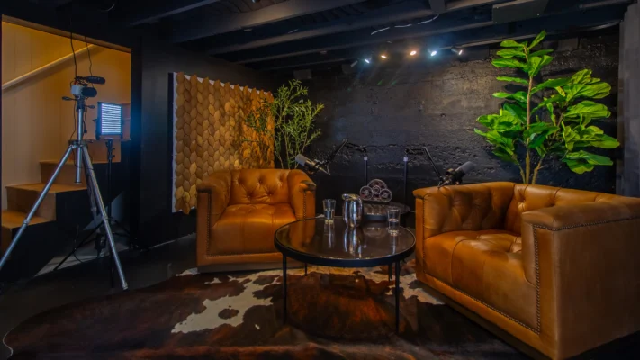 Podcast recording studio lounge area featuring two large, tufted brown leather armchairs, a round glass table, and two microphones on stands. The backdrop is a dark, textured wall with a fiddle leaf fig plant and a wooden scale pattern.