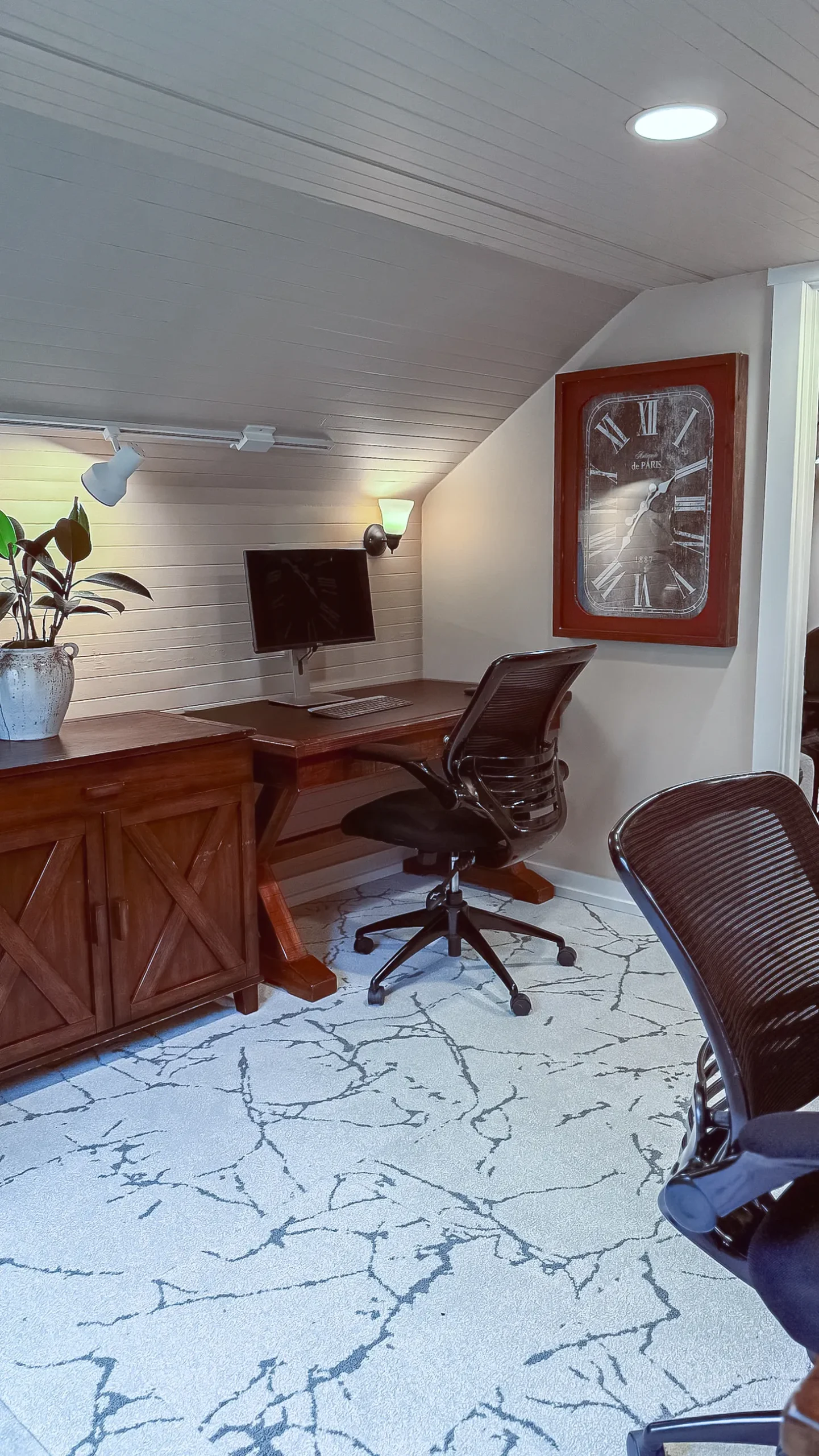 Vertical shot of a private office workspace with a dark wood desk, a black mesh chair, and a large wall clock with roman numerals against a sloped, shiplap ceiling.