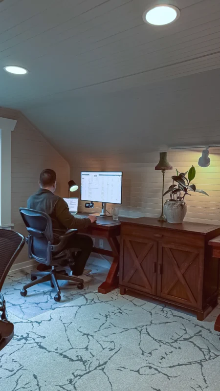 View of a man sitting at a dark wood desk in an attic office, working on a computer. A cabinet and potted plant are next to him.