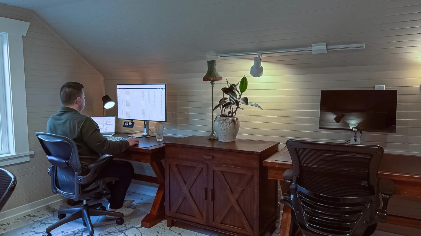 Horizontal view of two desks in a private office. A man is seated at the left desk working on a monitor; a plant and old-fashioned desk lamp sit between the two desks.