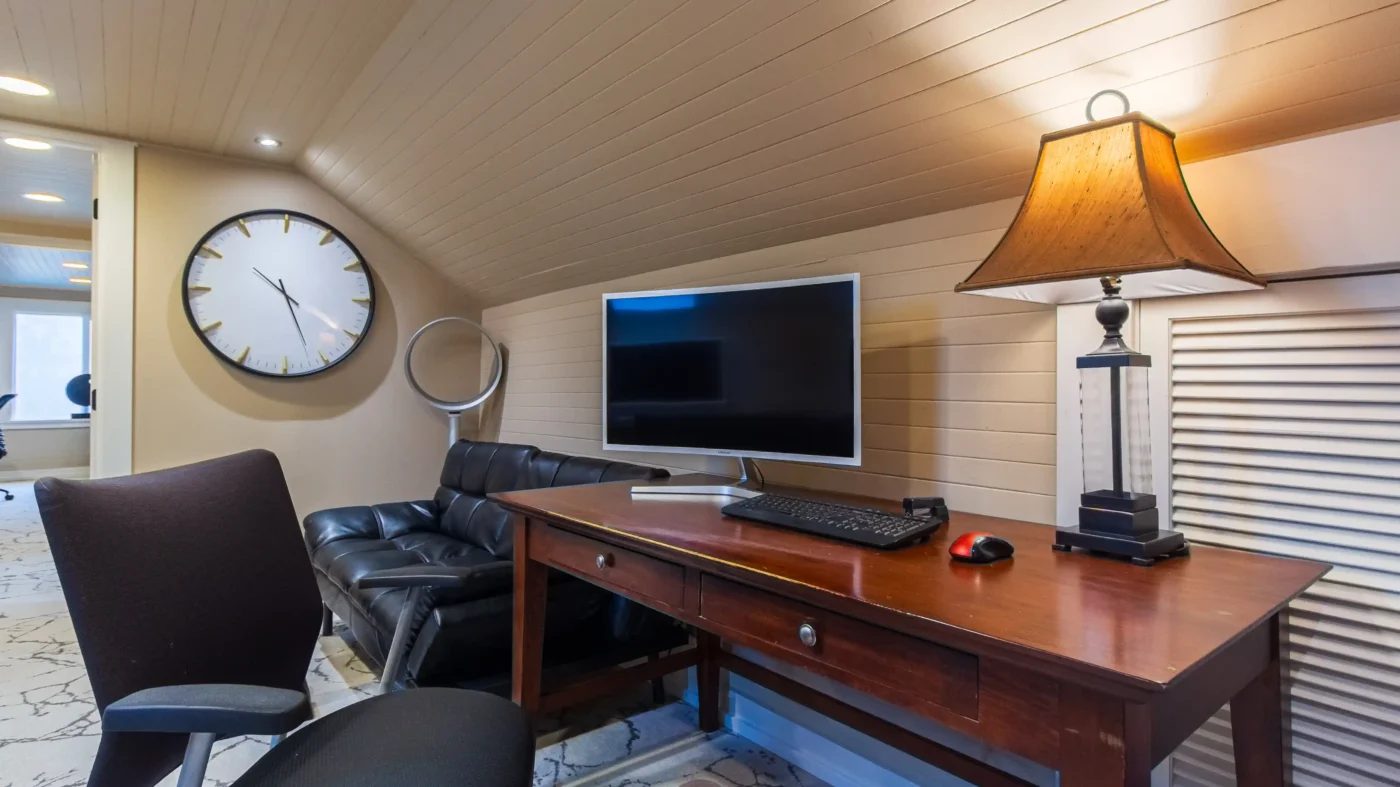 Private office with a large wall clock, black leather sofa, dark wood desk, and flat-screen monitor, set under a sloped, shiplap ceiling.