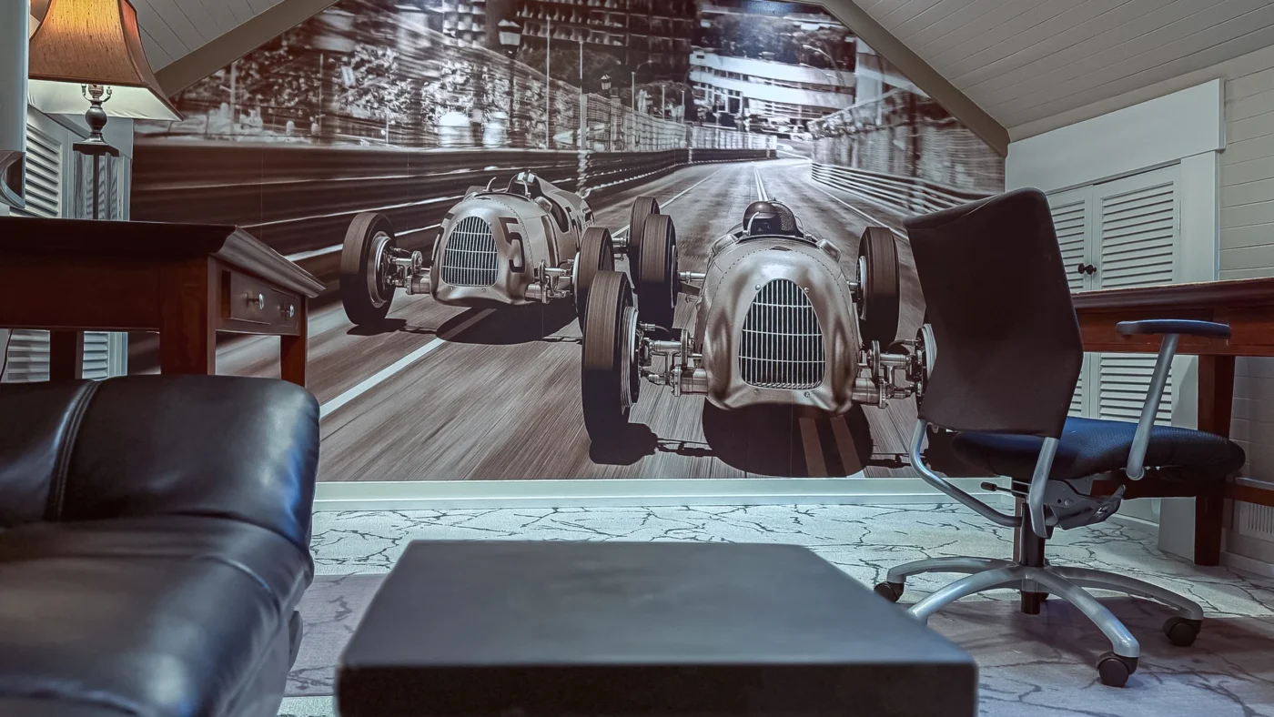 Wide view of a private office featuring a black-and-white mural of two vintage race cars on a sloped wall, with a black leather couch and an office chair in the foreground.